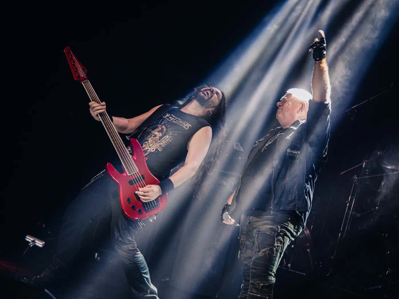 Fabio Carito playing a red bass next to Udo Dirkschneider, who raises his fist under the stage lights